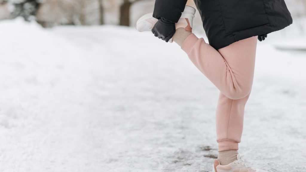 Person stretching in the snow, wearing winter attire and sneakers, in a serene outdoor setting.